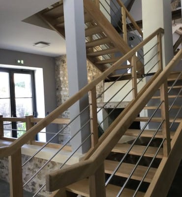 Escalier sur mesure en bois moderne avec rampe en tiges inox, sans contremarches, dans un intérieur lumineux avec mur en pierre apparente. Dreux